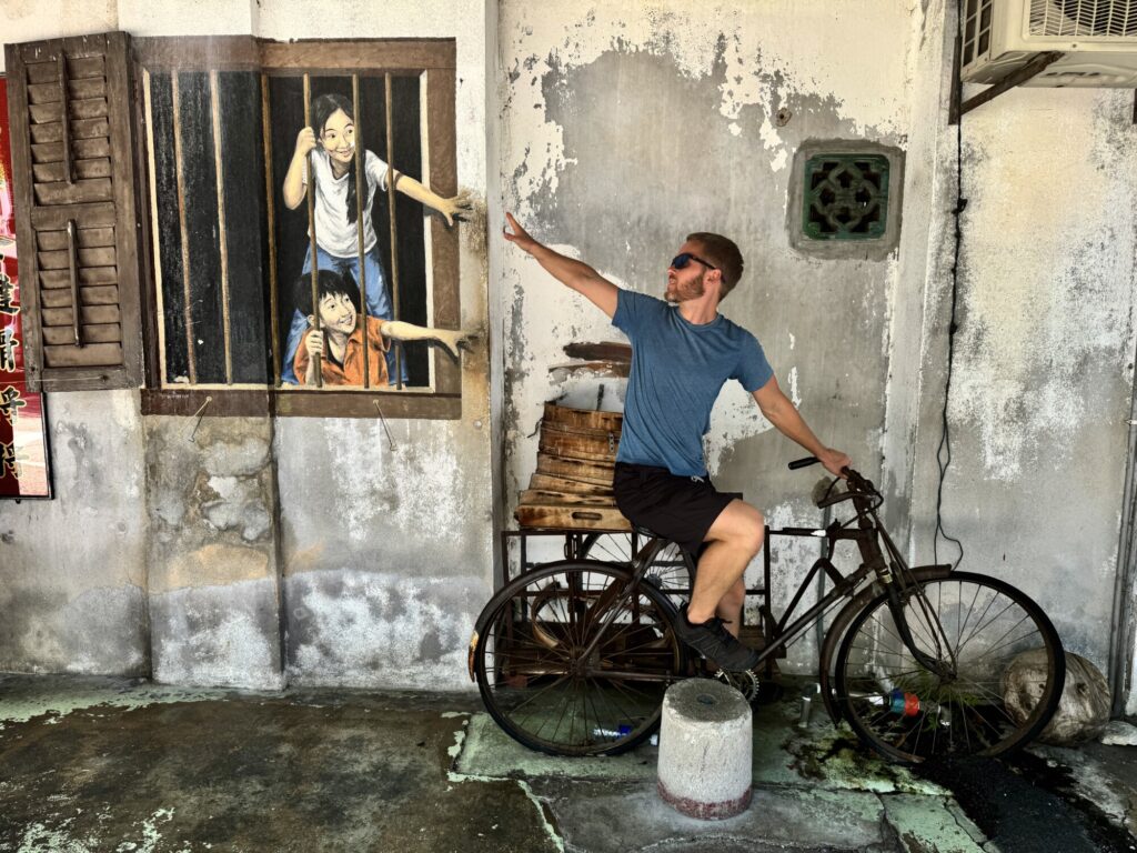 a man sitting on a bicycle with an interactive street mural of two kids reaching to the man on the bicycle