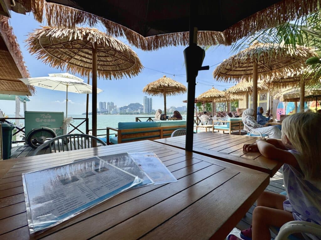 girl sitting at a table with palm frond umbrellas looking out at the ocean