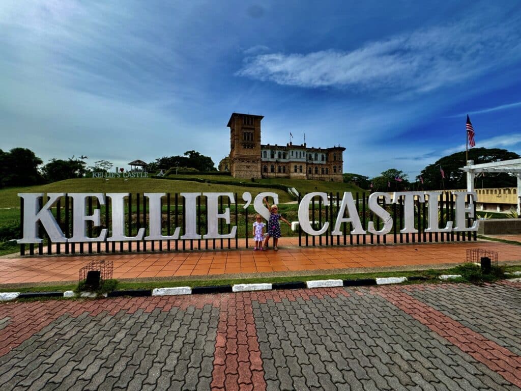 two girls standing in front of a sign of "Kellie's Castle" with the castle behind on a hill