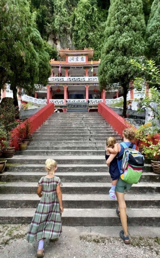 a woman carrying a child walking up the steps with a girl towards a temple amongst lush greenery