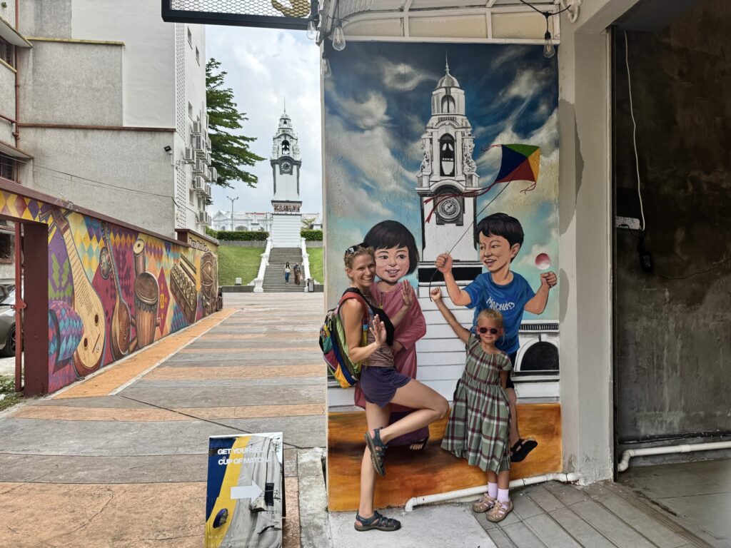 mother and a girl posing in front of a street mural with a clock tower depicted in the mural, with the real clock tower in the background