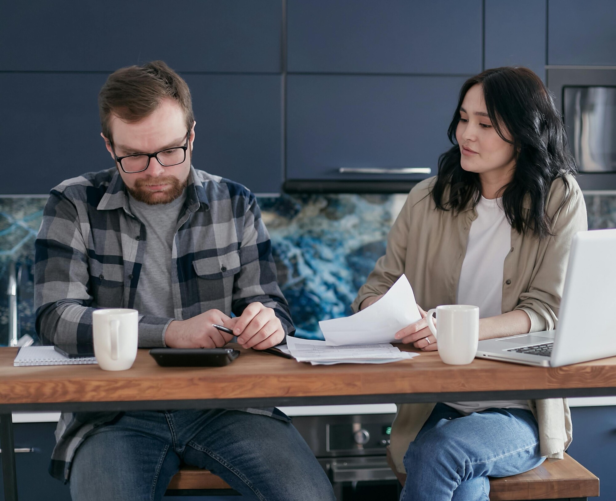 A couple discusses bills and finances using a laptop and calculator in their modern kitchen.