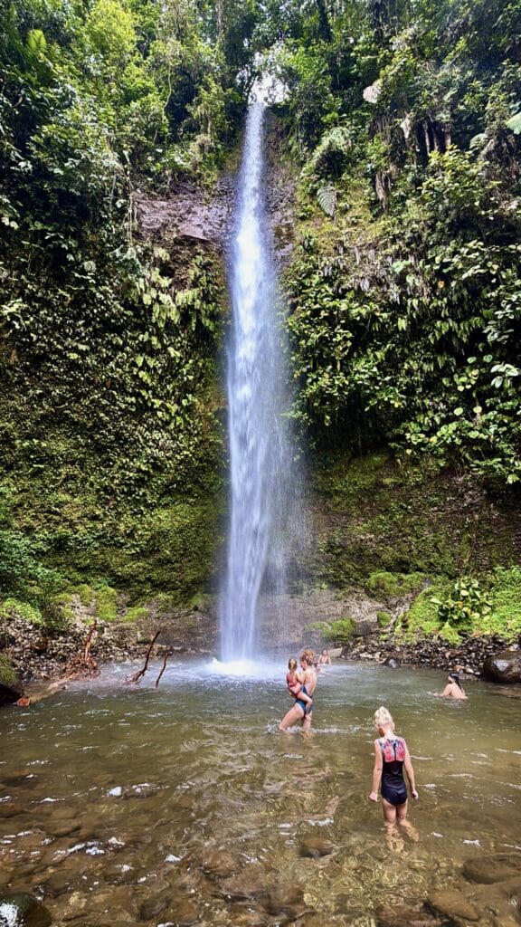 people standing and swimming in a shallow pool with a towering waterfall