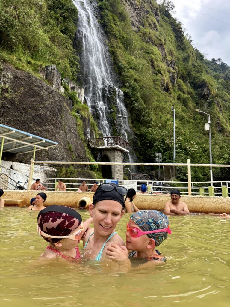 a mother and two girls with swim caps on and goggles swimming in natural hotsprings with a large waterfall in the background