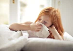 Small sick redhead kid with eyes closed cleaning nose with white tissue paper while lying alone in bed in light modern bedroom in daytime