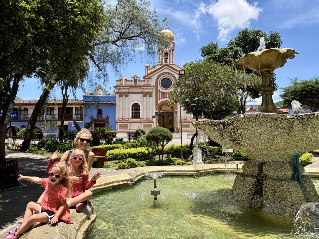 a woman and 2 girls sitting in a central square next to a fountain with a church in the background