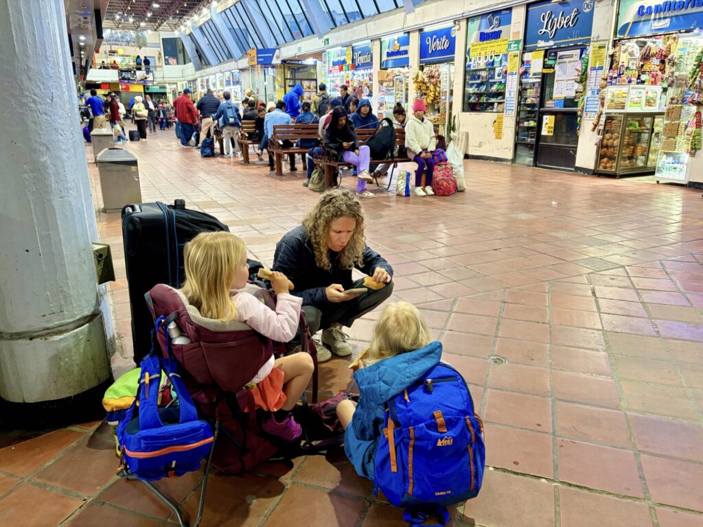 a mother and 2 girls sitting down at a bus station eating a snack