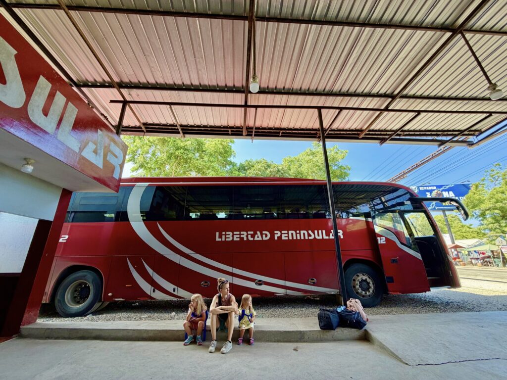 two girls and a mother sitting at a bus station with a bus in the background